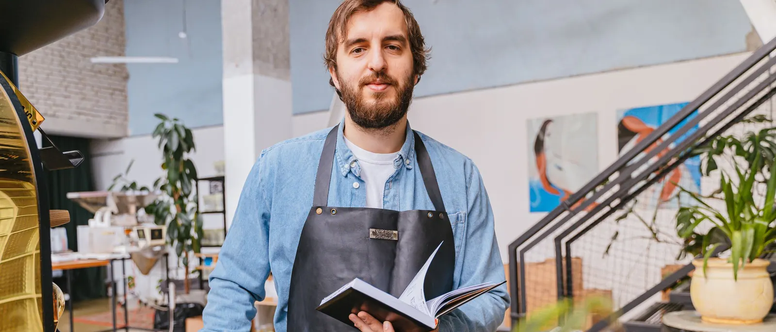 Un hombre con delantal sostiene un cuaderno en su tostadora de café.