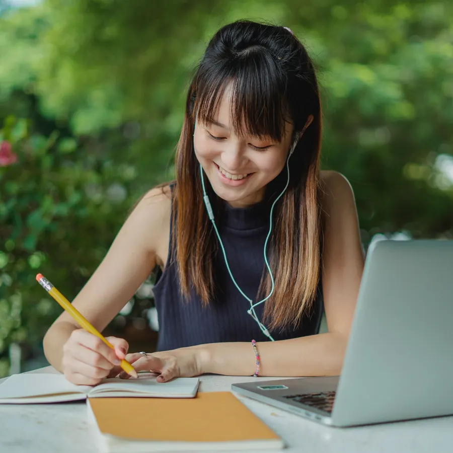 Una mujer toma notas con audífonos frente a su laptop, sonriendo.