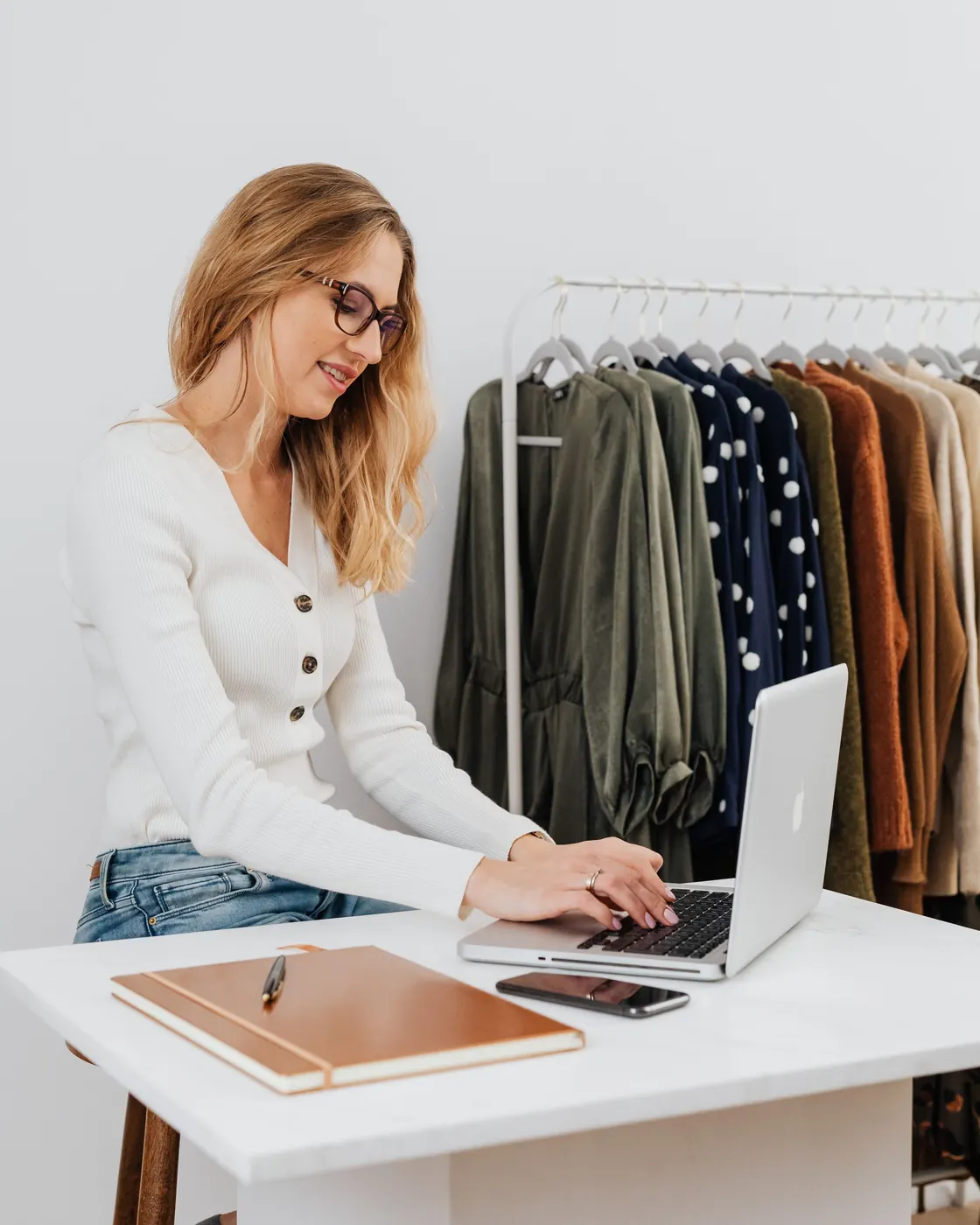 Una mujer trabaja en su laptop en su tienda de ropa, junto a un perchero.